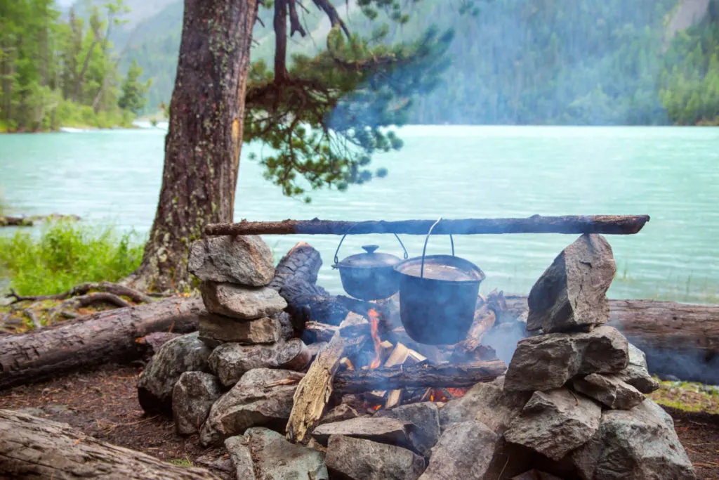 Lake in the background, foreground is a campfire with two pots suspended from a log on top of sev