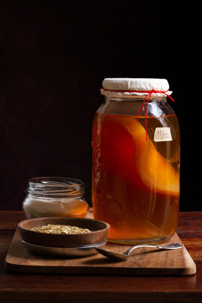 Tall jar of kombucha fermenting. Sugar and tea next to the jar on a cutting board. 