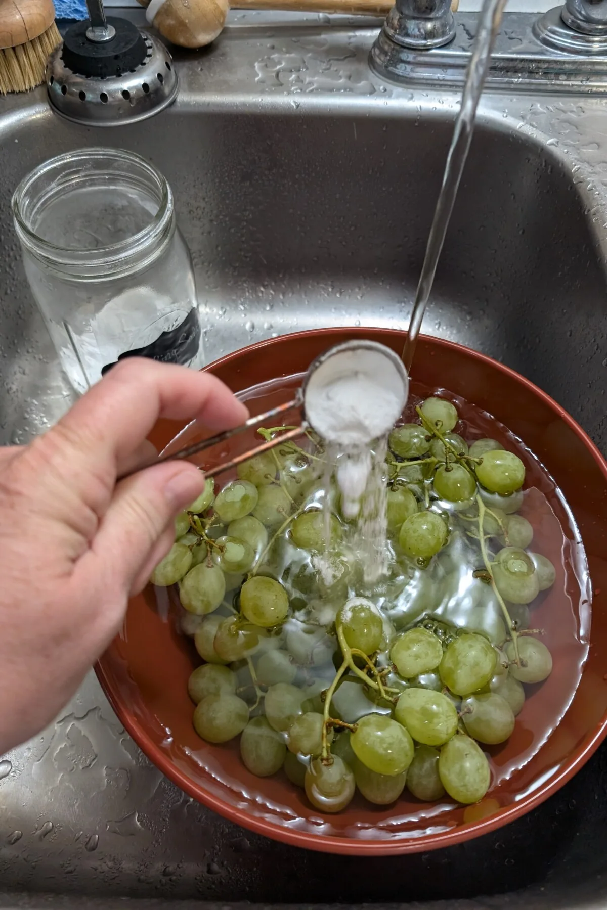 Hand pouring baking soda into a bowl filled with grapes and water